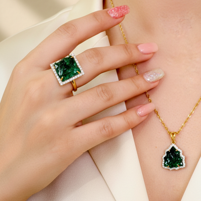 Person wearing a gold necklace with a green pendant and a matching ring on a plain background Elegant emerald and diamond gold ring on model – designer jewelry for women by Tresor Collection