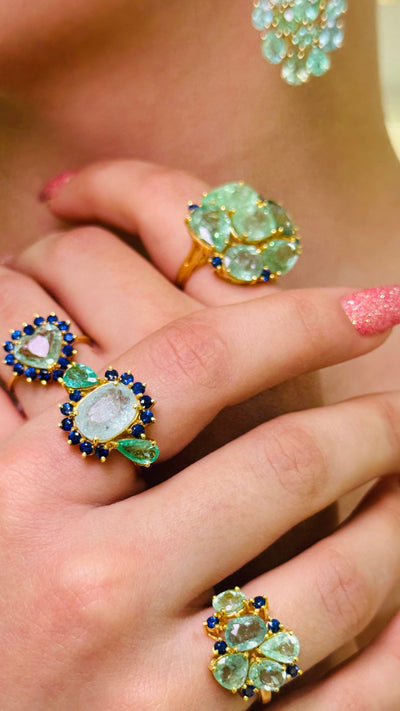 Close-up of hands wearing gold rings with green gemstones on a blurred background Elegant green and blue gemstone ring on hand – 18K gold designer jewelry for women by Tresor Collection Heart-shaped Paraiba tourmaline and blue sapphire halo ring on hand – bold luxe gemstone contrast