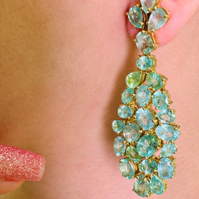 Close-up of a green gemstone earring on a person's ear with a blurred background. Close-up of 18K gold green gemstone chandelier earrings – handcrafted fine jewelry from Tresor Collection boutique