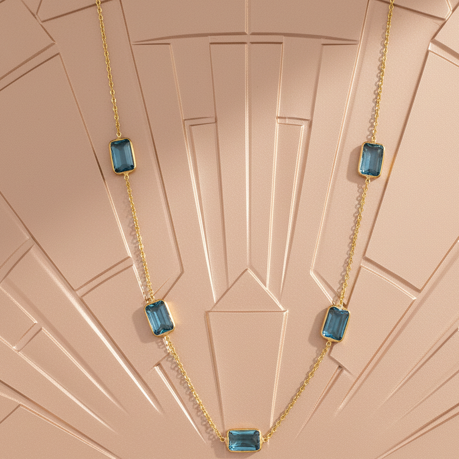 Gold necklace with blue gemstones on a patterned pink background Blue Topaz station necklace styled on silk fabric – premium handcrafted jewelry