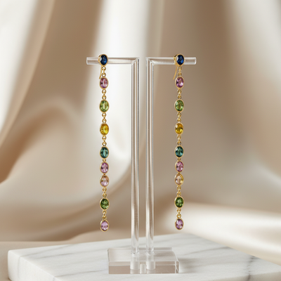 Pair of gemstone earrings on a clear stand with a soft beige background Multi-sapphire drop earrings in 18K gold displayed on jewelry stand – colorful luxury gemstone jewelry