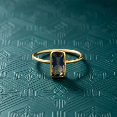 Gold ring with a blue gemstone on a textured green background Sapphire rectangular ring displayed on textured teal surface – premium handcrafted jewelry