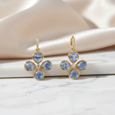 Gold earrings with blue gemstones on a soft fabric background Gold clover earrings with blue gemstones displayed on marble and satin – sophisticated jewelry styling