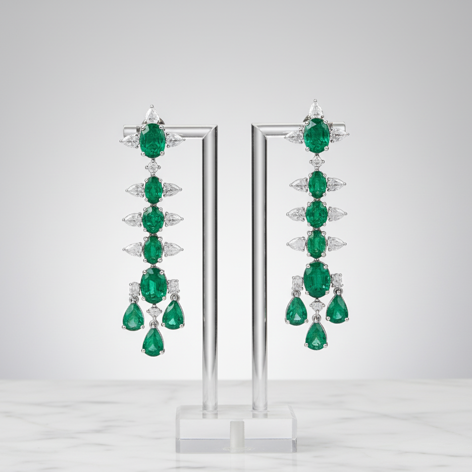 Pair of emerald earrings on a clear stand against a light gray background Long emerald and diamond drop earrings displayed on stand – handcrafted 18K gold jewelry