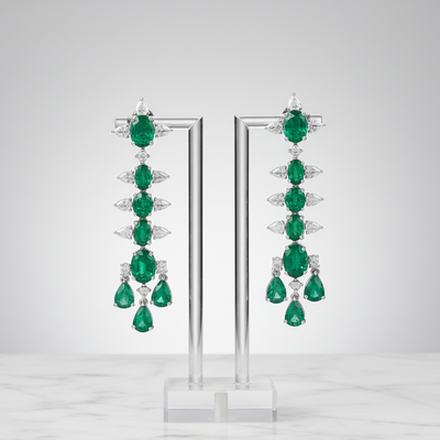 Pair of emerald earrings on a clear stand against a light gray background Long emerald and diamond drop earrings displayed on stand – handcrafted 18K gold jewelry