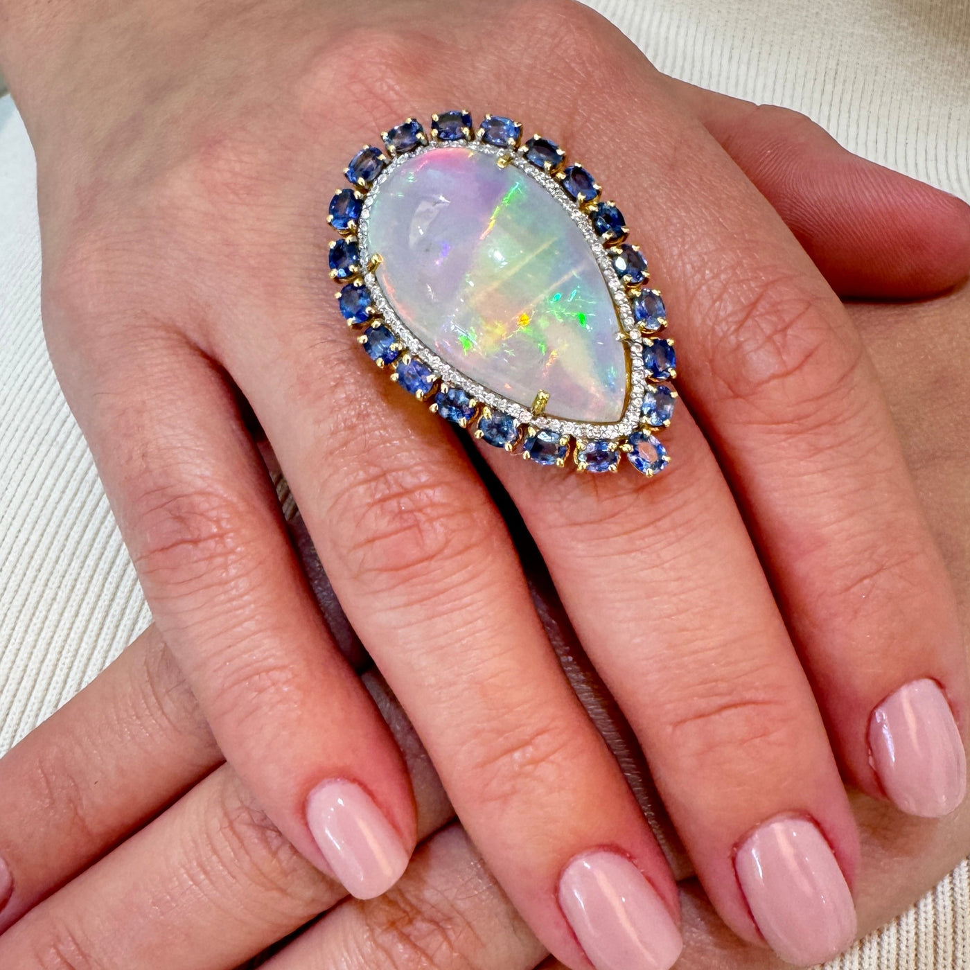 Ring with a large opal stone on a hand against a neutral background Close-up of Ethiopian opal and blue sapphire ring in 18K yellow gold – fine gemstone jewelry