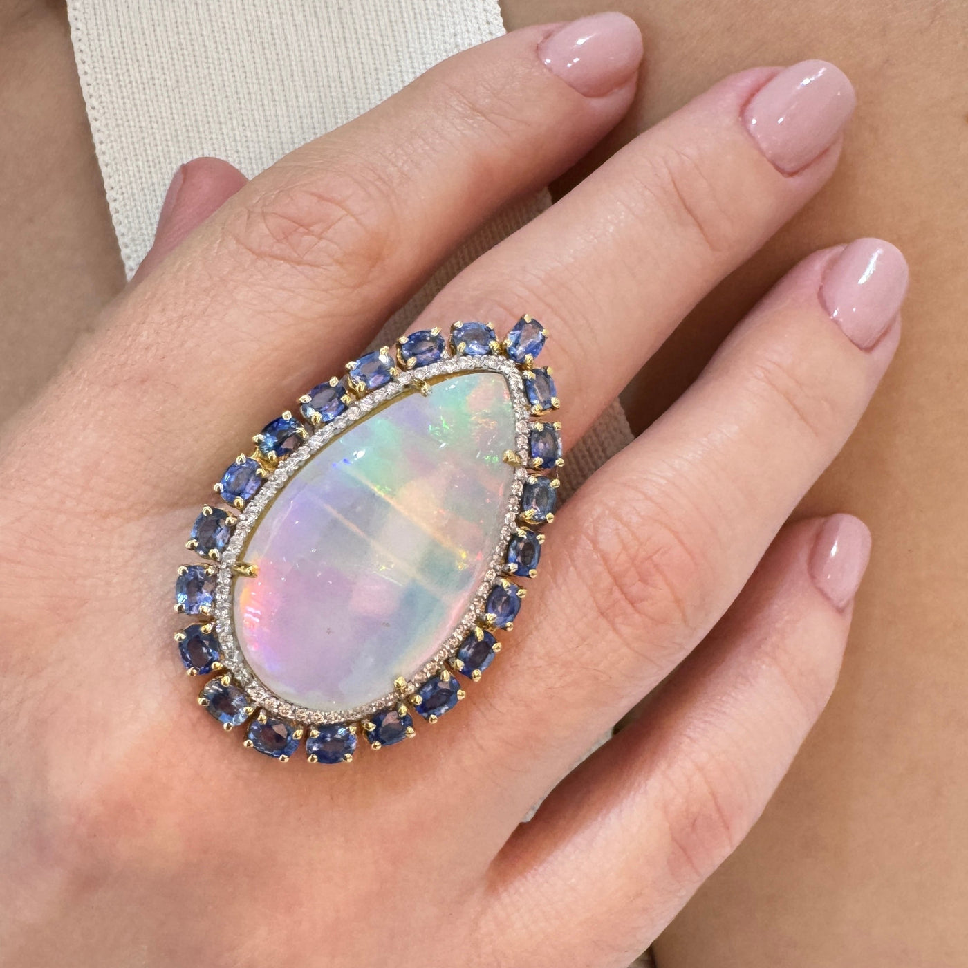 Close-up of a hand wearing an opal ring with blue gemstones on a light background Elegant Ethiopian opal and sapphire ring on model’s hand – designer jewelry for women by Tresor Collection