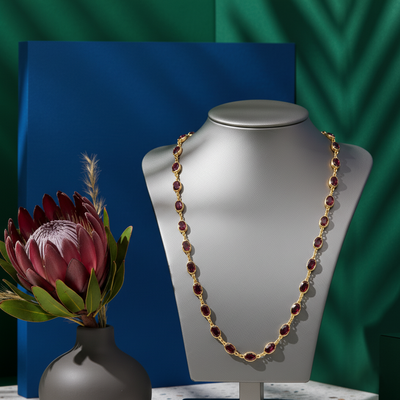 Gold and red beaded necklace on a mannequin bust with a flower arrangement in the foreground against a green and blue background. Close-up of 18K gold rhodolite necklace – handcrafted fine jewelry from Tresor Collection boutique