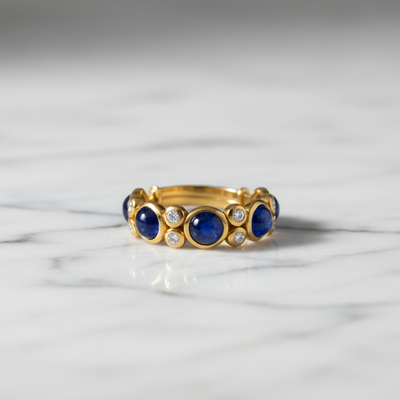 Gold ring with blue gemstones on a marble surface Close-up of 18K gold blue sapphire cabochon ring – handcrafted fine jewelry from Tresor Collection boutique