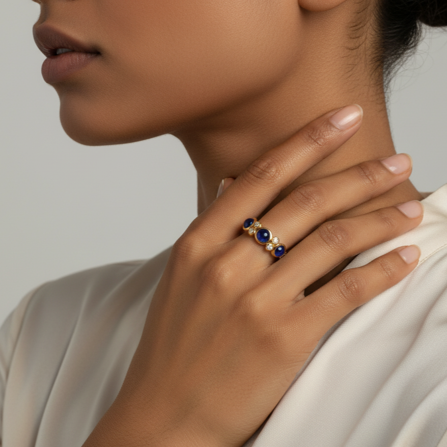 Close-up of a hand wearing a gold ring with blue stones, touching the neck against a neutral background. Elegant blue sapphire and diamond ring on model – designer gold jewelry for women by Tresor Collection