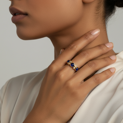 Close-up of a hand wearing a gold ring with blue stones, touching the neck against a neutral background. Elegant blue sapphire and diamond ring on model – designer gold jewelry for women by Tresor Collection